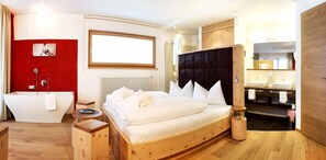 Une chambre à coucher moderne avec un grand lit, une tête de lit en bois, une armoire intégrée, une salle de bain séparée avec une baignoire et un mur d’accent rouge.