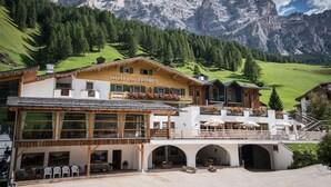 A hotel with a wooden facade, large windows, and a balcony with flowers. It is surrounded by green hills and mountains.