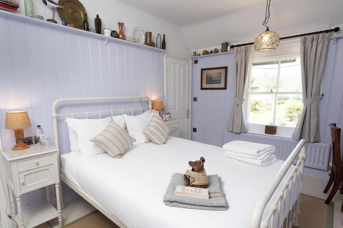 A neatly arranged bedroom with a bed, bedside tables, a window with curtains, and a teddy bear on a blanket.