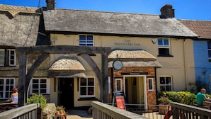 The Smugglers Inn mit traditionellem Strohdach, einer sichtbaren Uhr und einem Schild für Hall & Woodhouse.