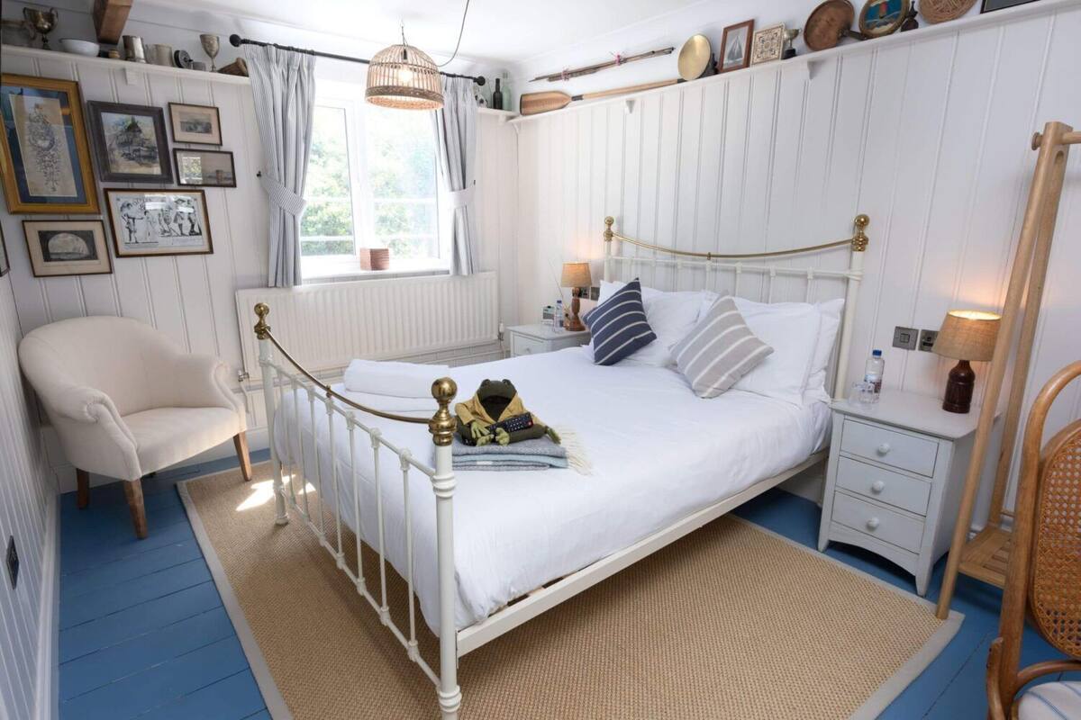 A bedroom with a white metal bed frame, a white upholstered chair, a bedside table with a lamp, and a window with curtains.