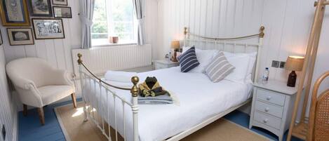 A bedroom with a white metal bed frame, a white upholstered chair, a bedside table with a lamp, and a window with curtains.