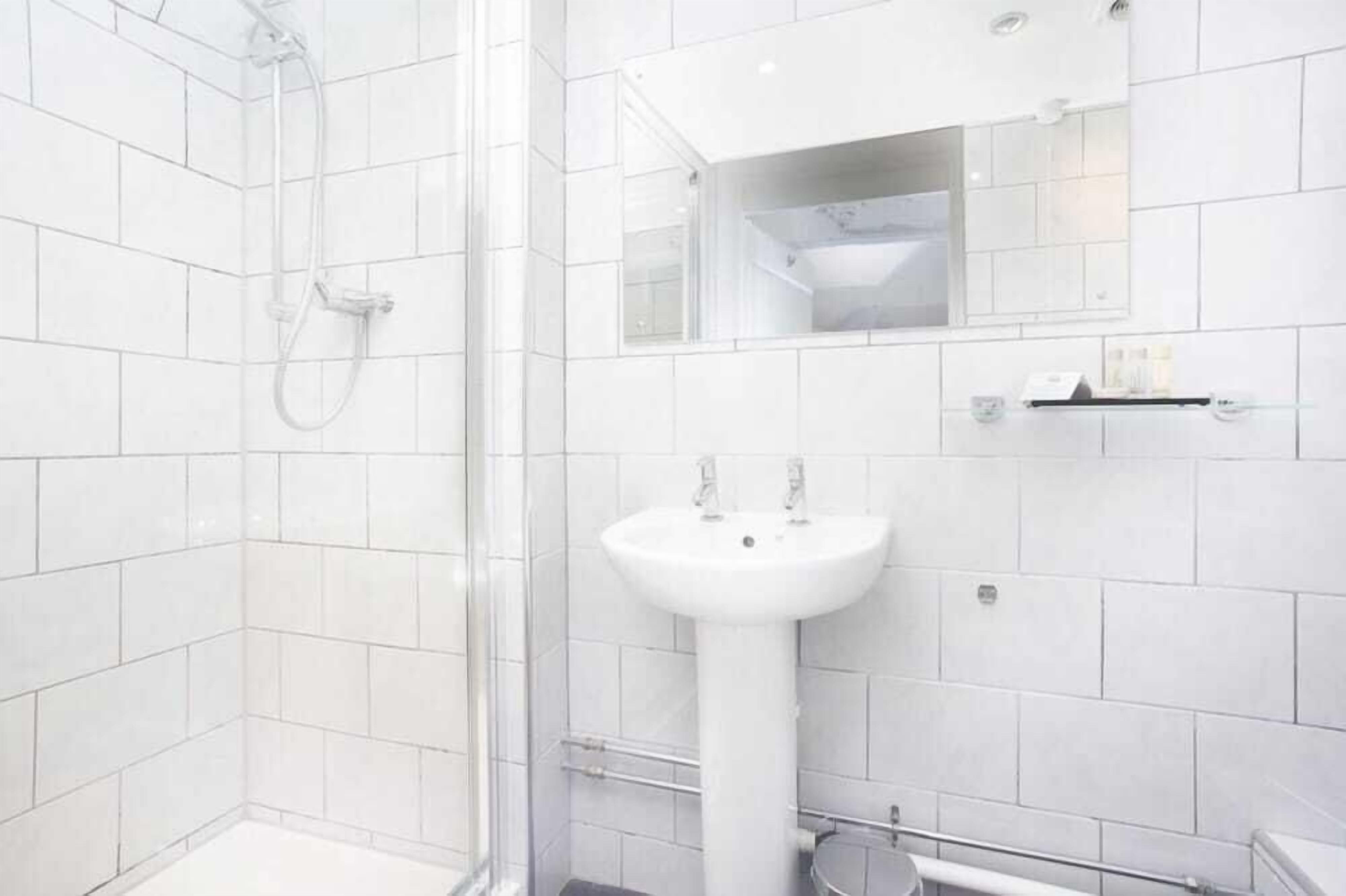 A modern bathroom with white tiles, a glass shower enclosure, a pedestal sink, and a mirror above the sink.