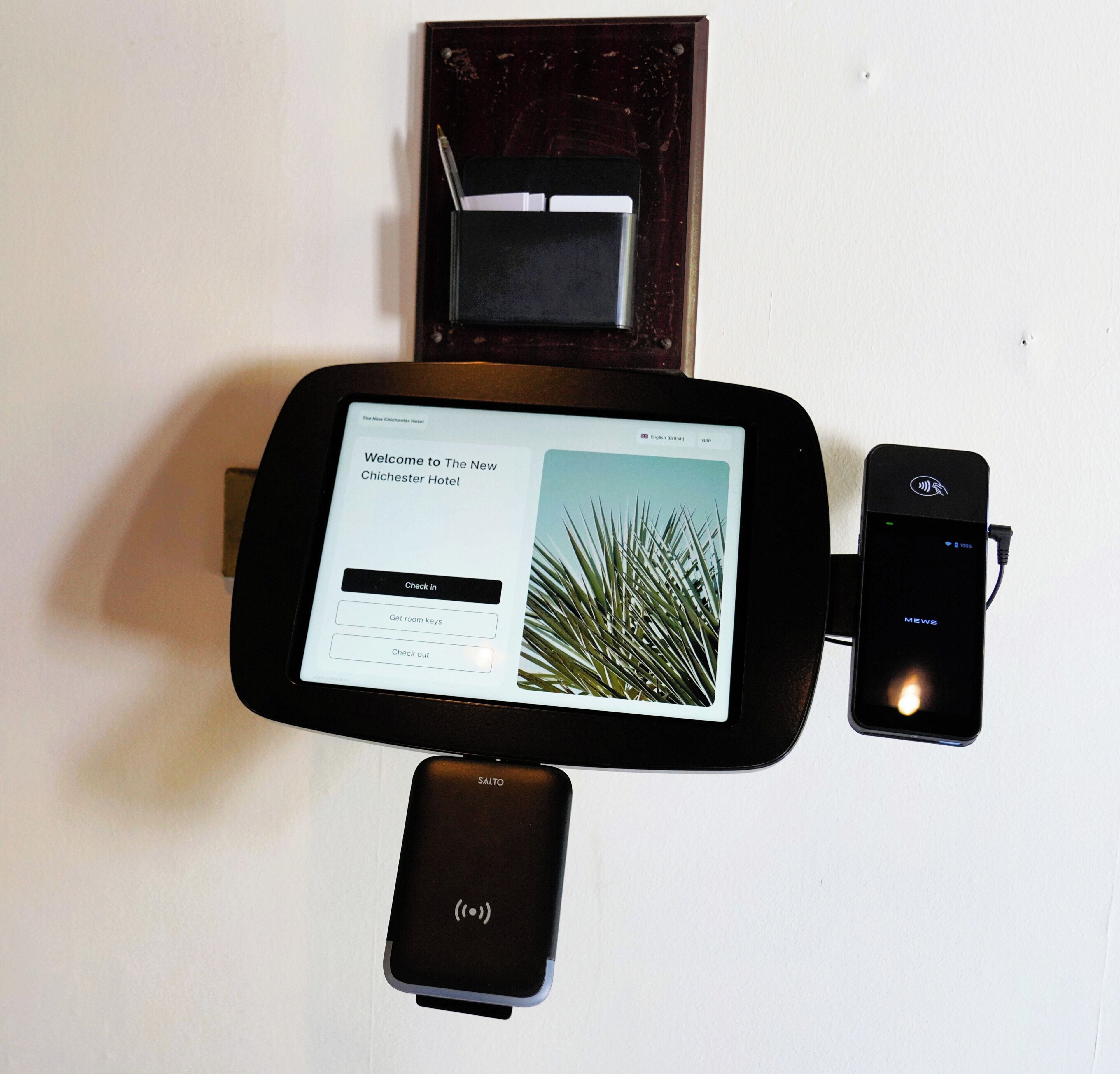 Quiosco digital de registro de entrada con pantalla mostrando un mensaje de bienvenida para The New Chichester Hotel, opciones para registrarse, llaves de habitación y salida.