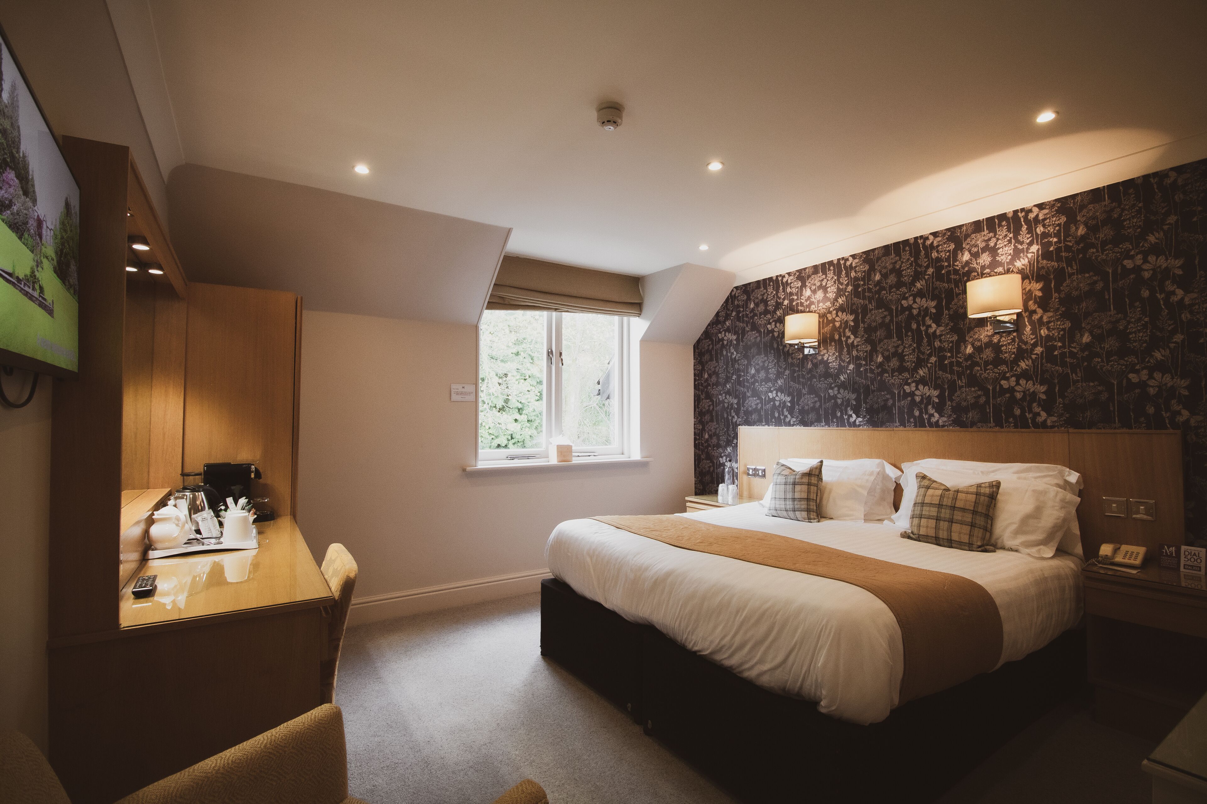 A hotel room with a bed, bedside tables, a desk, a chair, a window, and a wall-mounted TV.