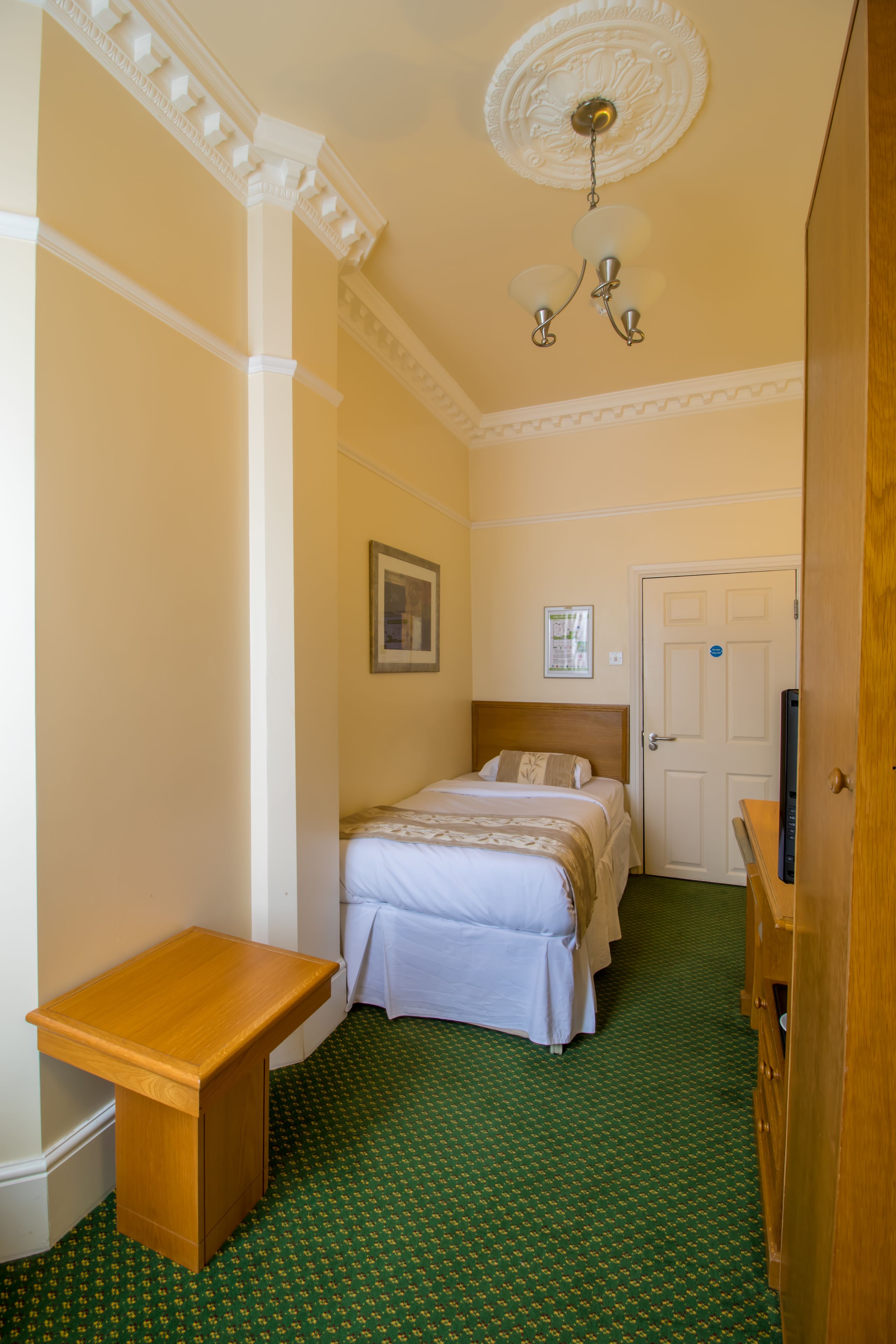 A hotel room with a bed, a wooden nightstand, a green patterned carpet, a white door, and a ceiling light fixture.