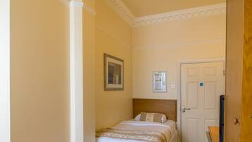 A hotel room with a bed, a wooden nightstand, a green patterned carpet, a white door, and a ceiling light fixture.