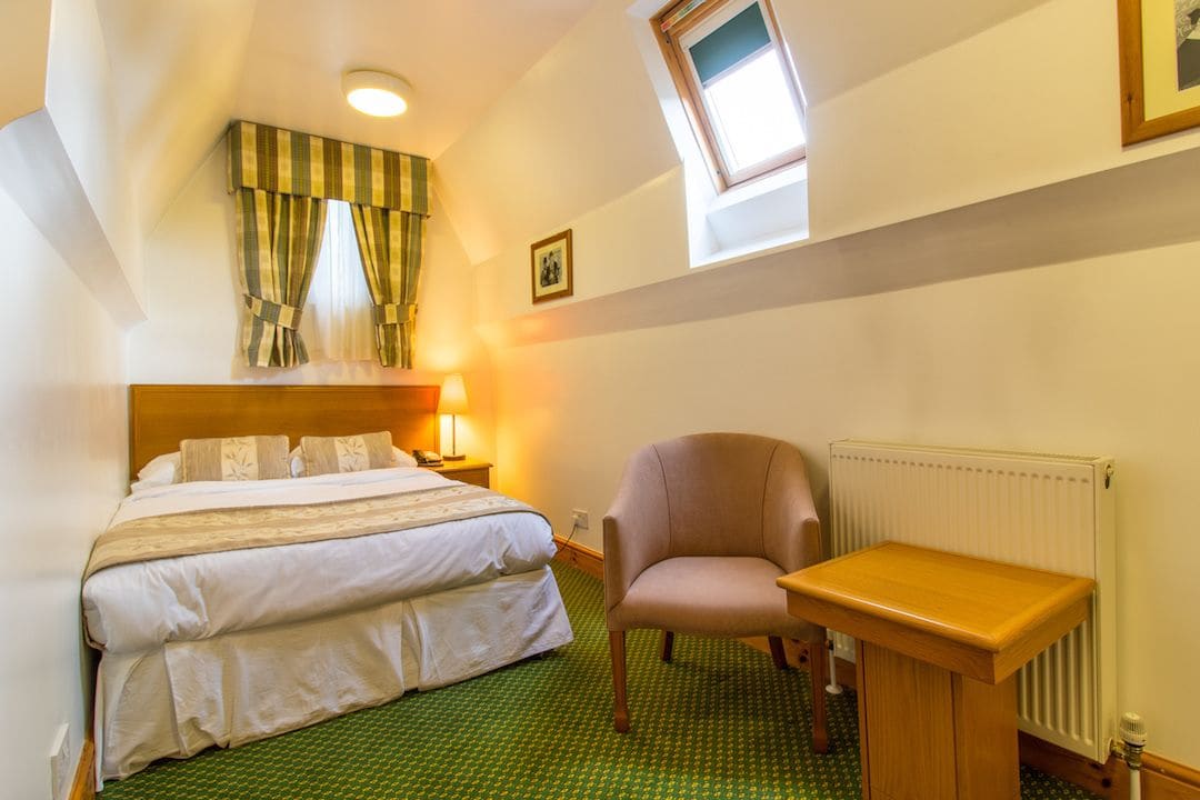 A hotel room with a double bed, a chair, a small table, a radiator, and a window with curtains.