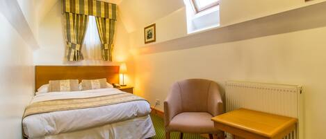 A hotel room with a double bed, a chair, a small table, a radiator, and a window with curtains.