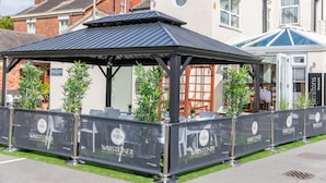 A covered outdoor seating area with a black roof, surrounded by barriers with 'Warsteiner' branding, and adjacent to a building with a blue window and a sign for 'HAMILTON Restaurant'. - A Park View Hotel (Wolverhampton)