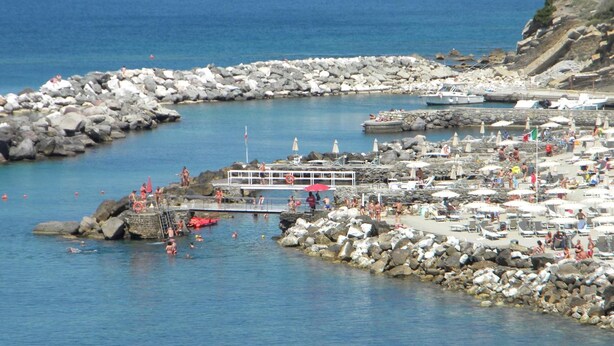 Zona costera con terreno rocoso, un muelle y una playa con sombrillas y gente.