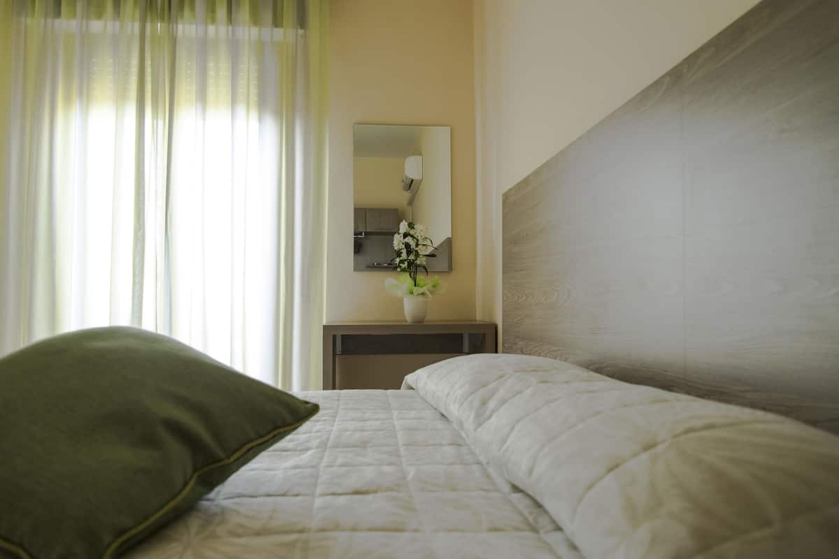 Een netjes opgemaakt bed met een groen kussen, een nachtkastje met een bloemenvaas, en een spiegel aan de muur.