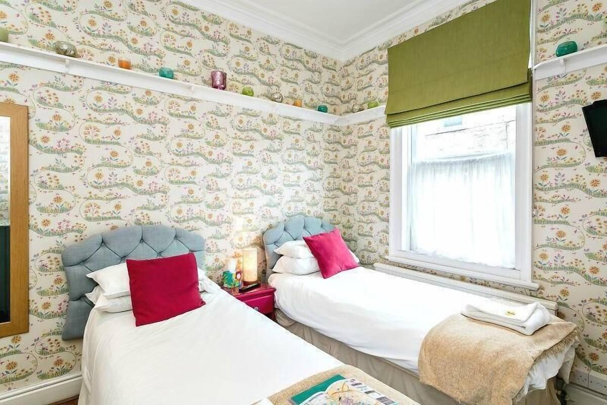 Une chambre à coucher avec un papier peint à motifs floraux, deux lits avec des têtes de lit, une table de chevet, une fenêtre avec des rideaux verts et une étagère ornée d’objets décoratifs.
