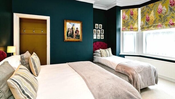 Un dormitorio con dos camas, una pared verde azulado oscuro, un valance de ventana con motivos florales y un cuadro en la pared.
