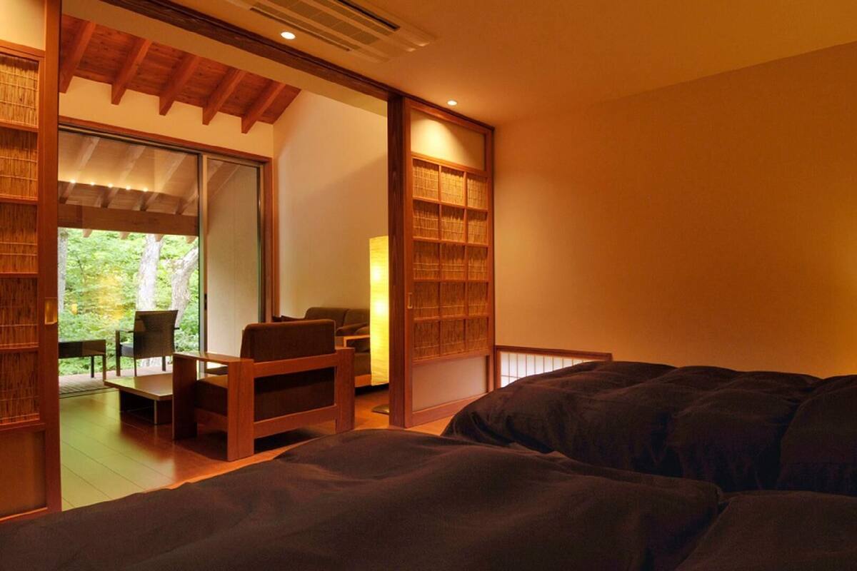 Um quarto com cama, escrivaninha e cadeiras, porta deslizante para uma varanda e uma janela grande.