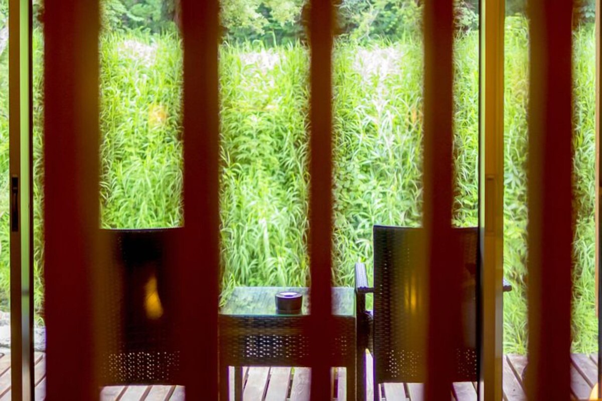 Une vue à travers une fenêtre avec des stores verticaux, une terrasse en bois et une table avec une chaise.