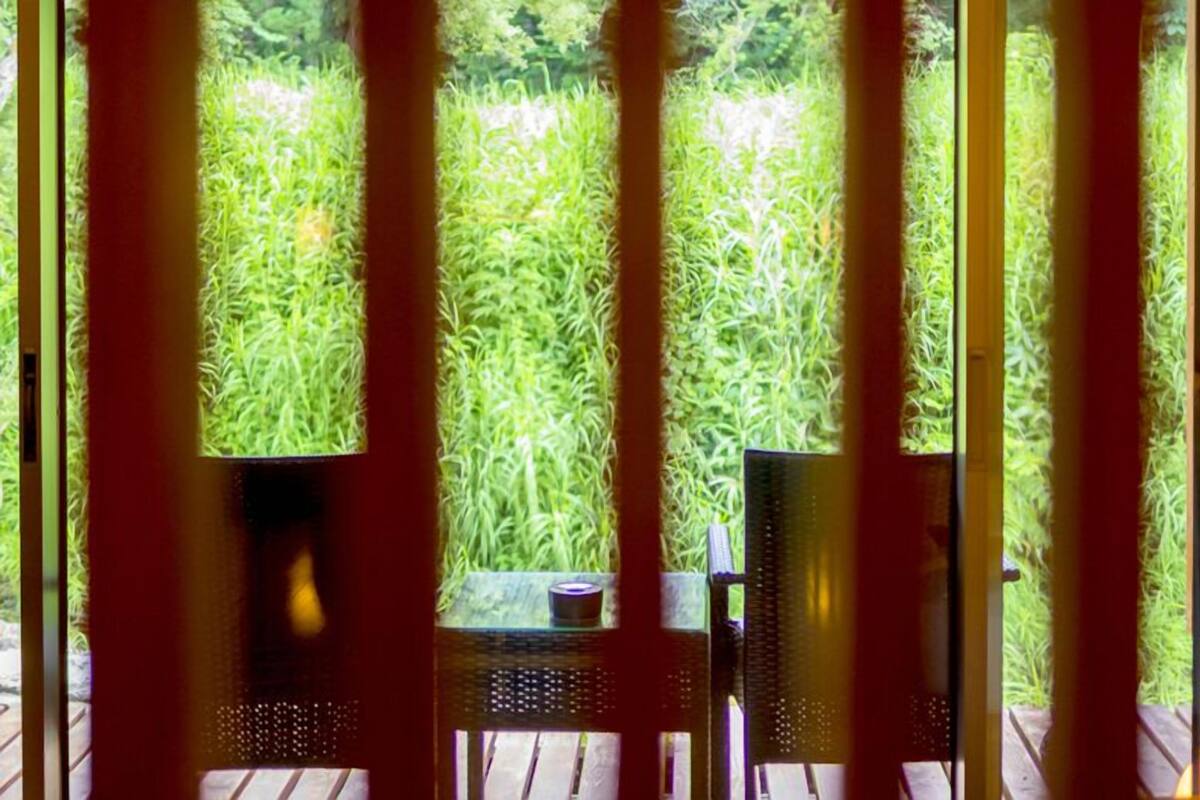 Vista pela janela, com persianas verticais, um deck de madeira e uma mesa com uma cadeira.