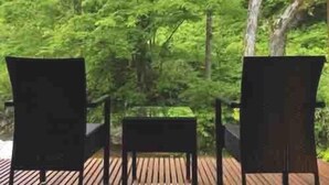 Un área de estar al aire libre con dos sillas y una mesa, rodeada por una terraza de madera y abundante follaje verde.