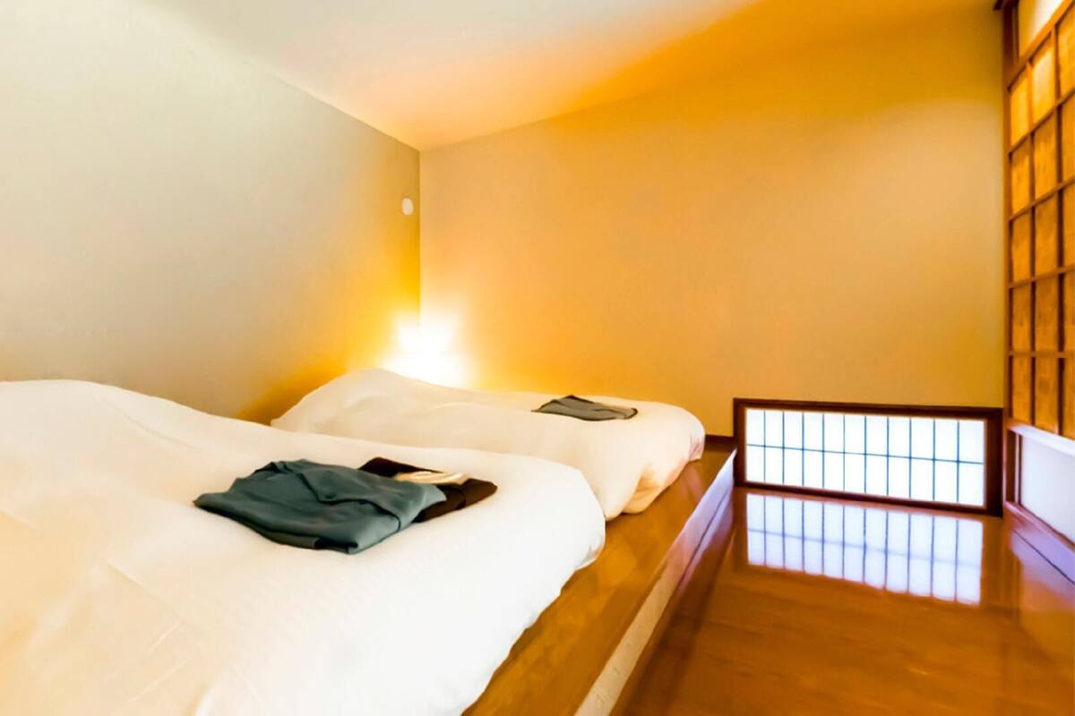 Quarto com duas camas, mesa de cabeceira, luminária fixada na parede e vista pela janela com moldura de madeira.