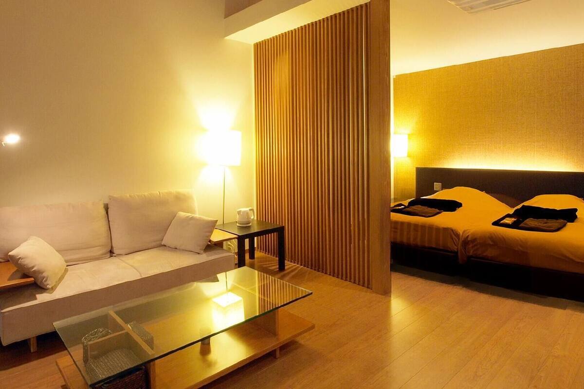 Quarto de hotel com sofá, mesa de centro de vidro, mesa de cabeceira com abajur, cama com almofadas e painel de madeira vertical.