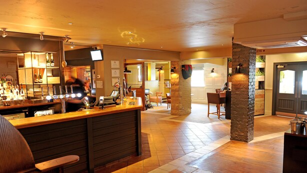 A bar area with a counter, stools, and a television screen. There are tables and chairs in the background. - Premier Inn York North West (York)