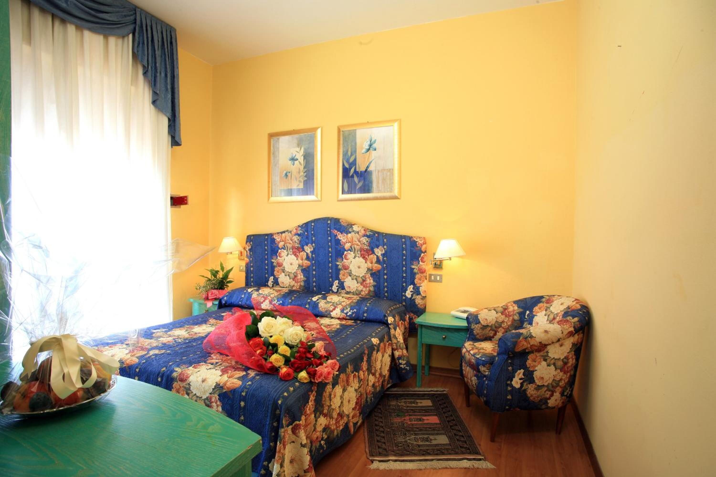 A bedroom with a floral bedspread, a bedside table with a lamp, a chair, and two framed pictures on the wall.
