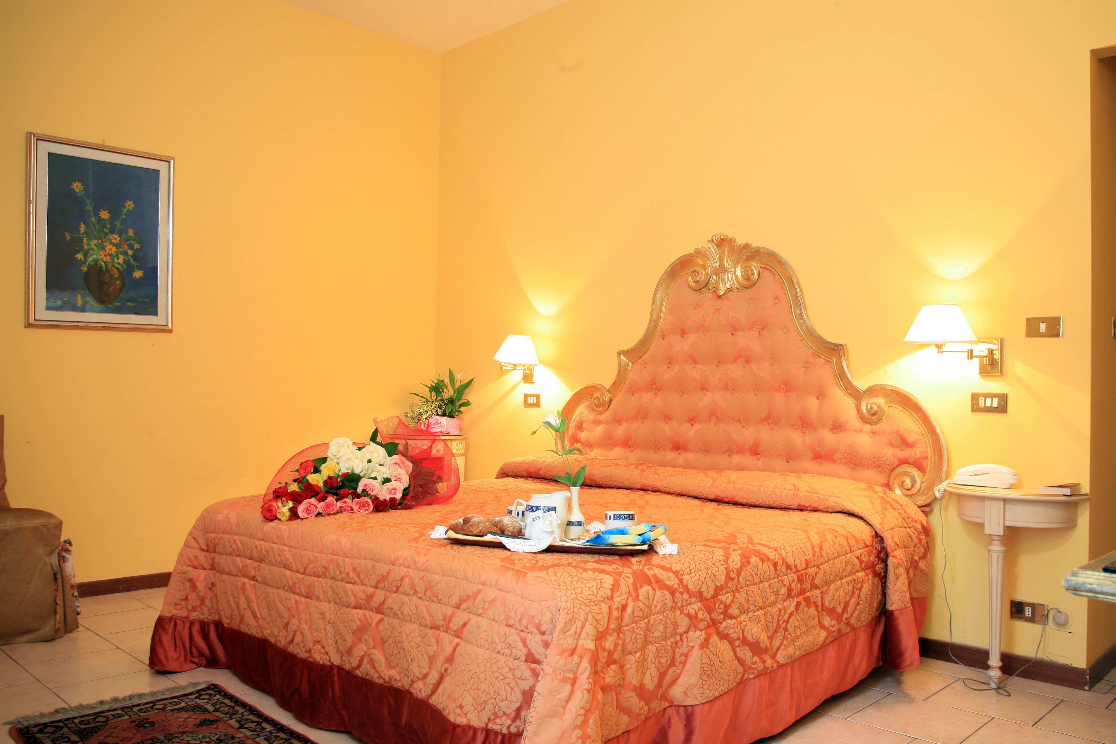 A hotel room with a large bed, a bedside table with a lamp, a vase with flowers, and a painting on the wall.