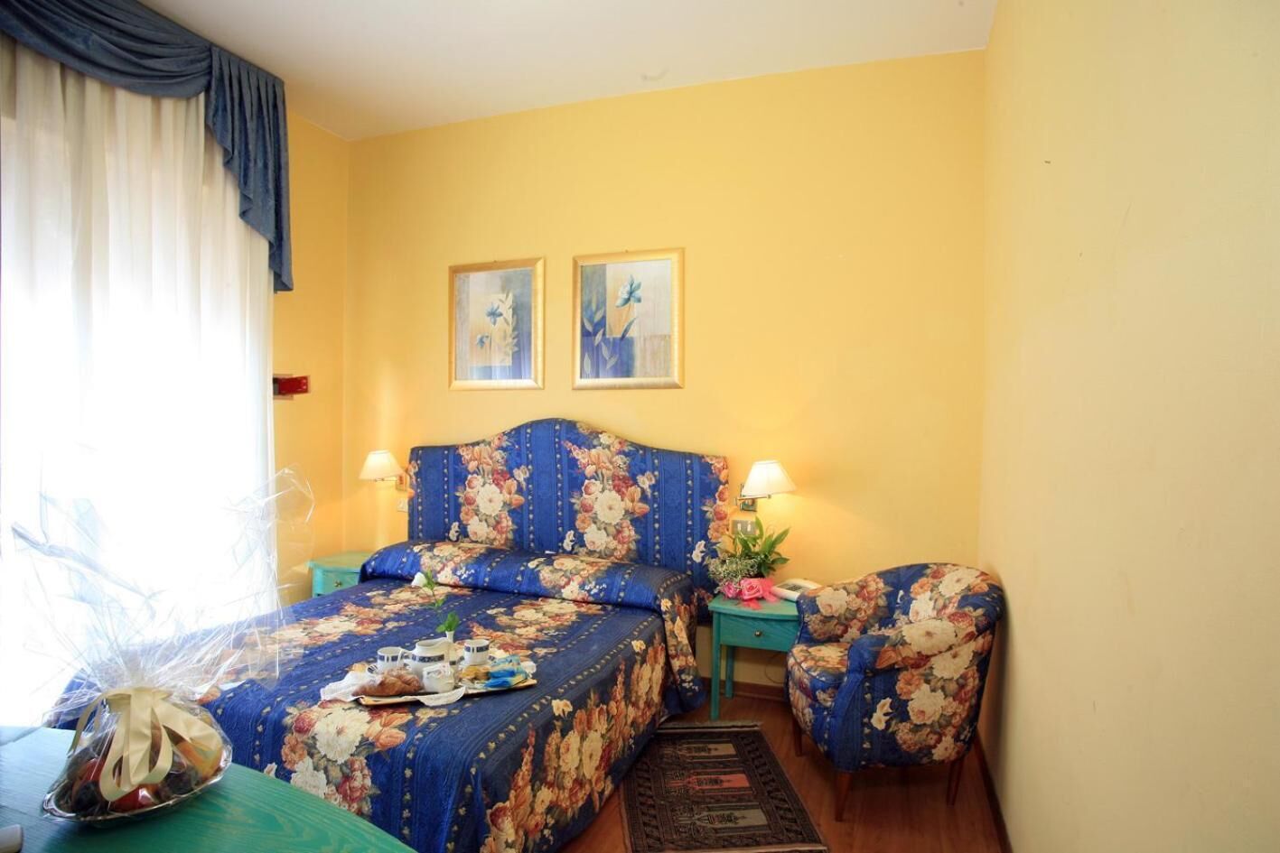 A bedroom with a floral bedspread, two bedside tables with lamps, a chair, and a small table with a decorative item.