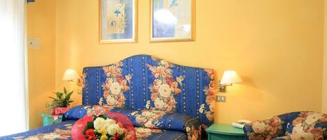 A room with a floral bedspread, a headboard, two wall-mounted lamps, a bedside table with a lamp, a chair, and a bouquet of flowers on the bed.