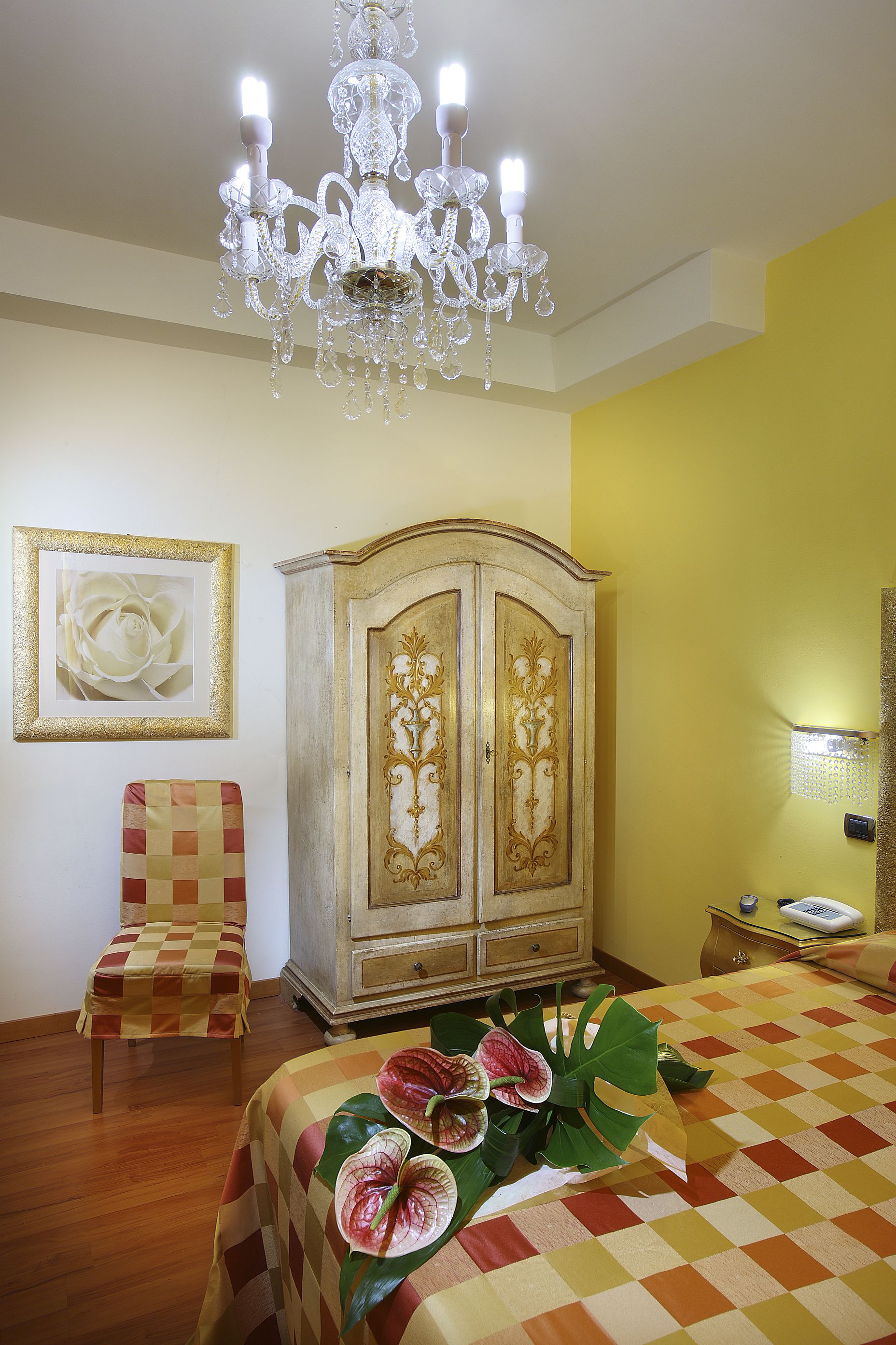 A hotel room with a chandelier, a large cabinet, a checkered chair, a bed with a floral bedspread, and a framed picture on the wall.
