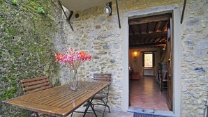 A rustic stone wall with a wooden table and chairs, a vase with flowers, and a doorway leading to an interior space. - Casa Vacanze Le Muse Sillico (Pieve Fosciana)