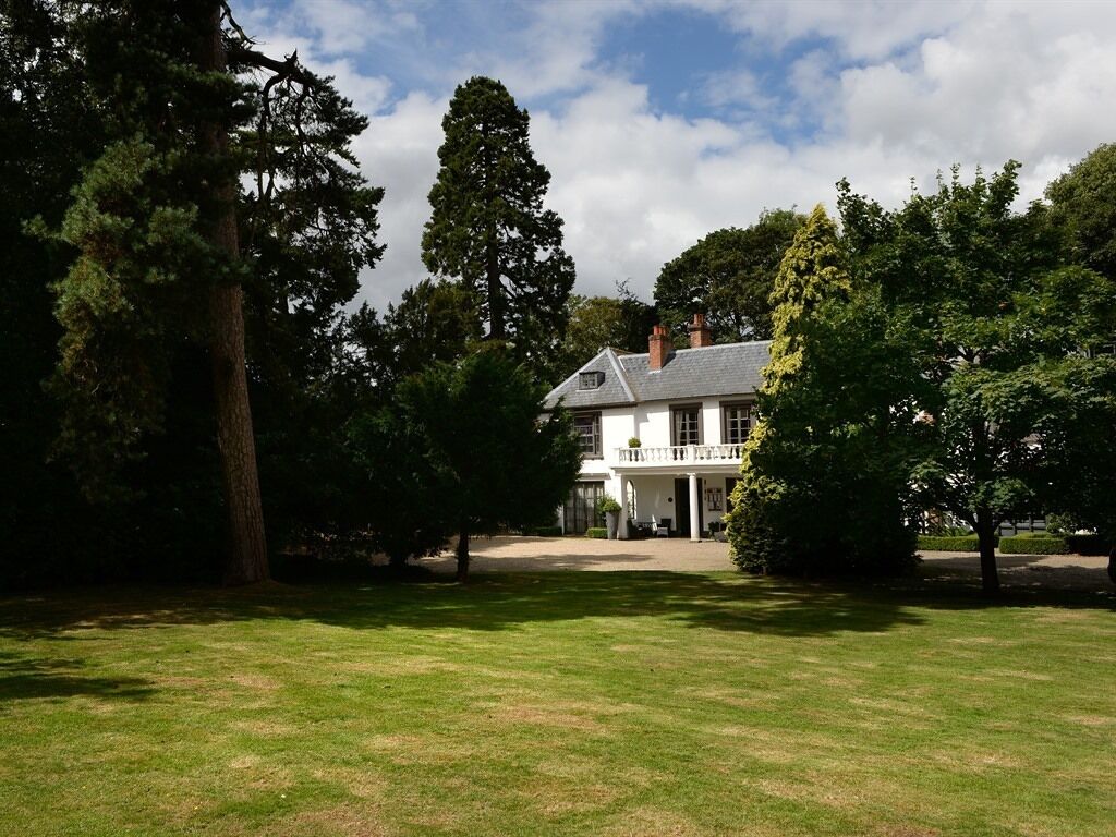 A large, white house with a grey roof, surrounded by a well-maintained lawn and tall trees.