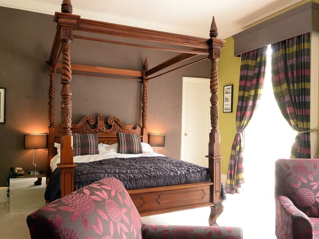 Ein Bett mit Baldachin, dessen dunkles Holzgestell aufwendige Schnitzereien aufweist, ein Ohrensessel mit Blumenmuster sowie der Blick auf eine Tür und Vorhänge.