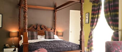 Ein Bett mit Baldachin, dessen dunkles Holzgestell aufwendige Schnitzereien aufweist, ein Ohrensessel mit Blumenmuster sowie der Blick auf eine Tür und Vorhänge.