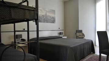 Una habitación de hotel con literas, un escritorio con lámpara, una silla, una mesita con lámpara y un cuadro de paisaje urbano en la pared.