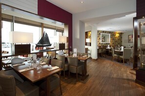 Comedor de hotel con mesas y sillas de madera, y un ventanal con vista a un velero.