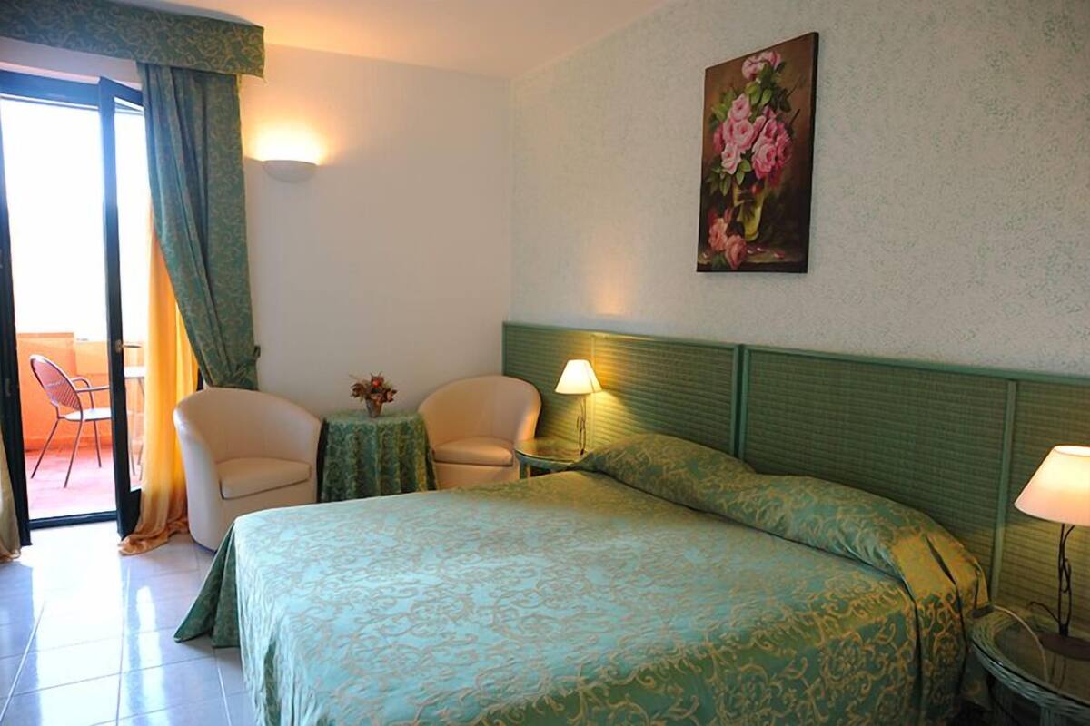 Ett hotellrum med en säng, två stolar, ett litet bord med en vas med blommor och en tavla på väggen.