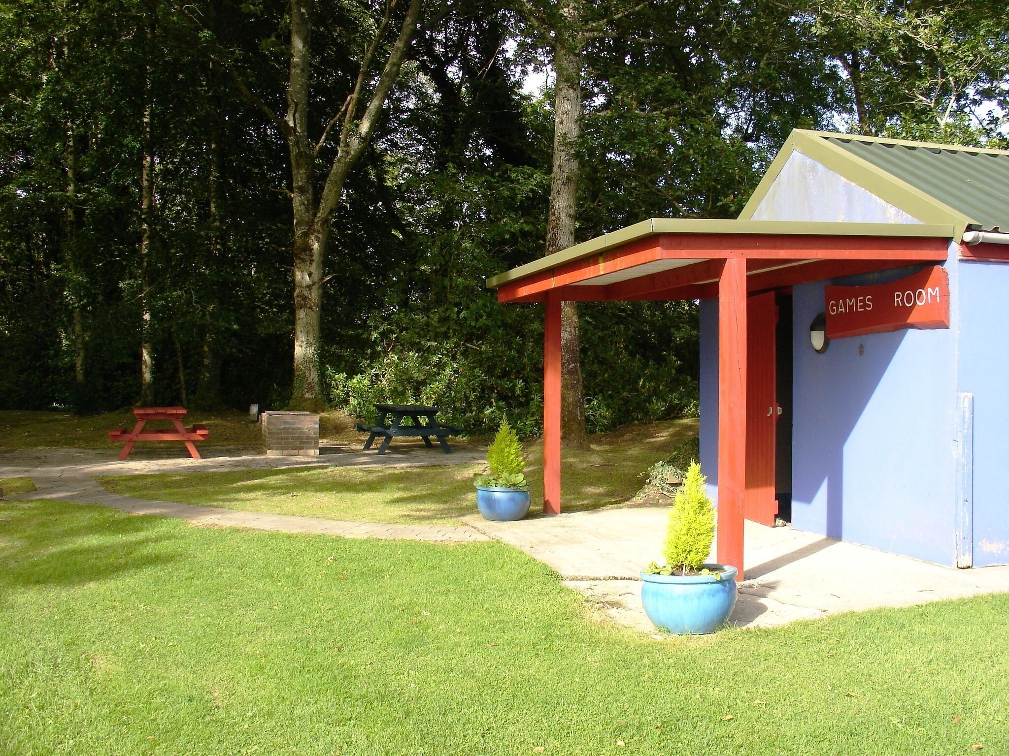 Een klein gebouw met de tekst 'GAMES ROOM', voorzien van een rood dak en blauwe muren, omringd door een gazon met picknicktafels en plantenpotten.