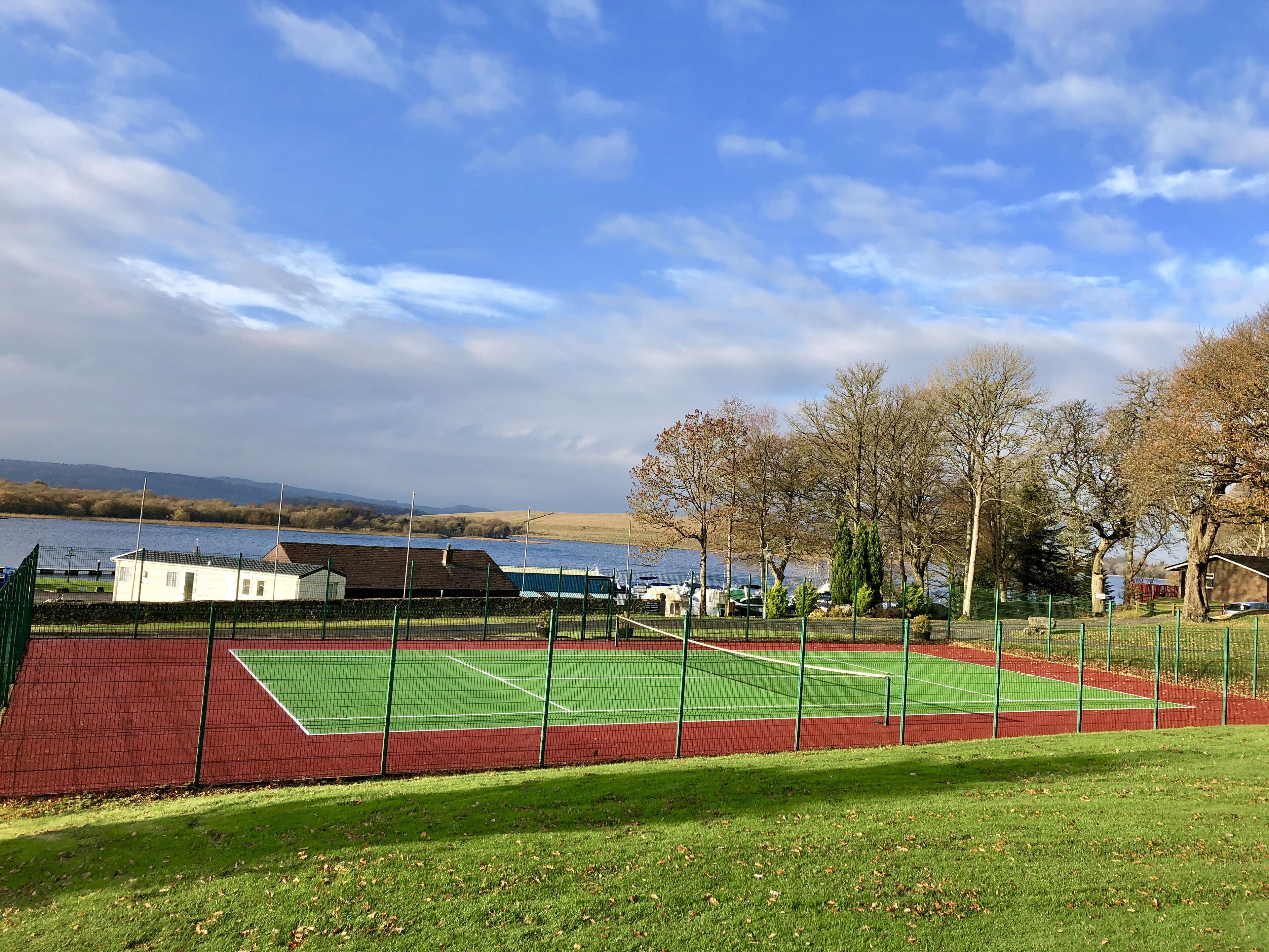 A tennis court with a net, surrounded by a green fence, near a body of water and trees.