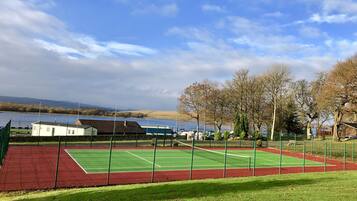 A tennis court with a net, surrounded by a green fence, near a body of water and trees.