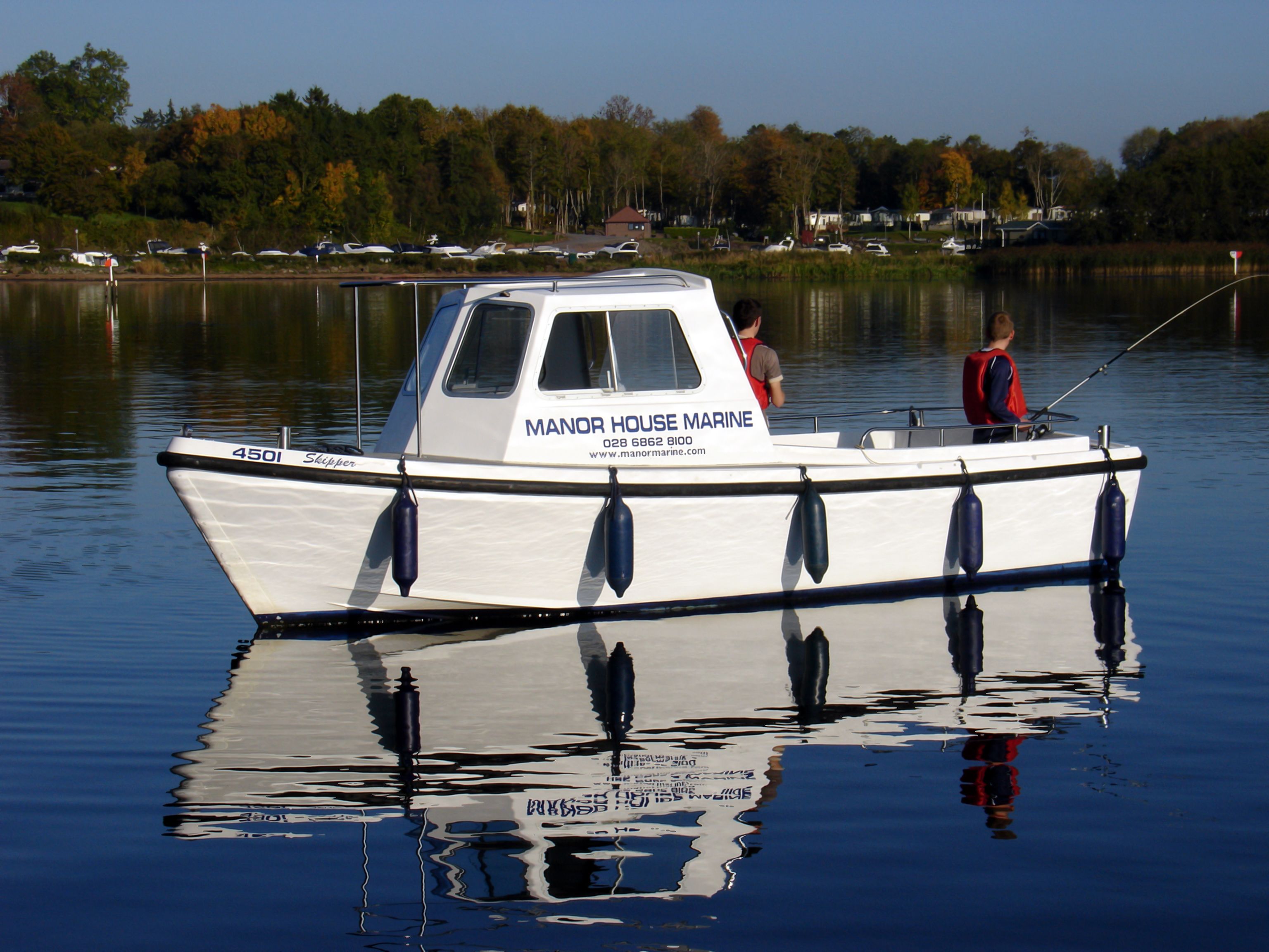 Een witte boot met de naam "Manor House Marine" op de zijkant, afgemeerd in kalm water.