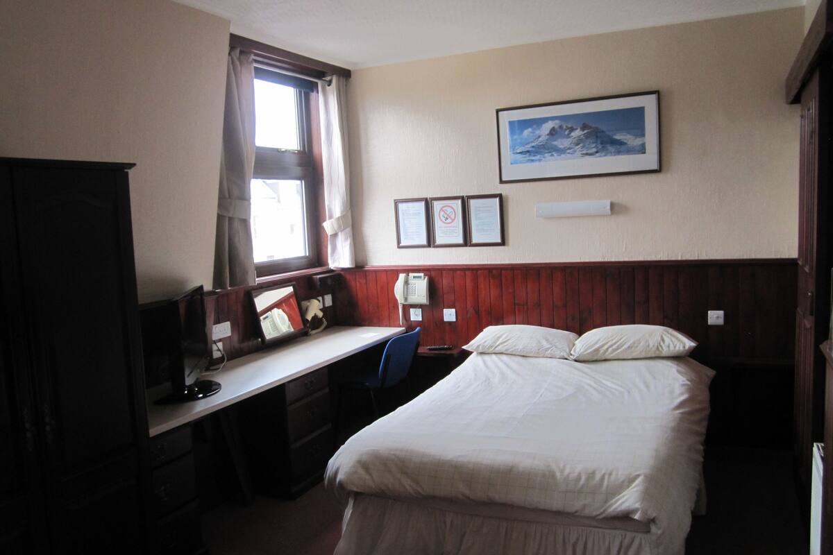 Ein Schlafzimmer mit Bett, Schreibtisch, Stuhl, einem Fenster mit Vorhängen, einem gerahmten Bild an der Wand und einem Schrank.