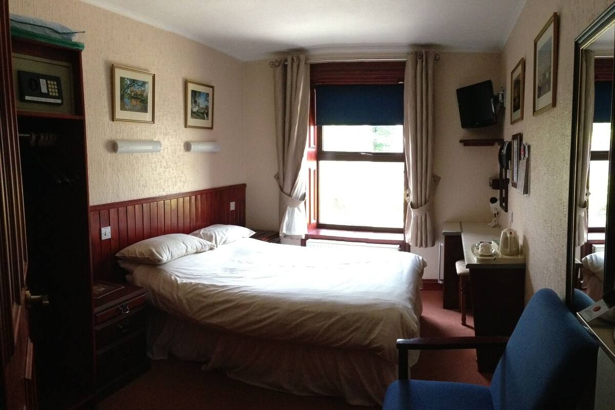 Ein Hotelzimmer mit Bett, Sessel, Schreibtisch, Fernseher und einem Fenster mit Vorhängen.
