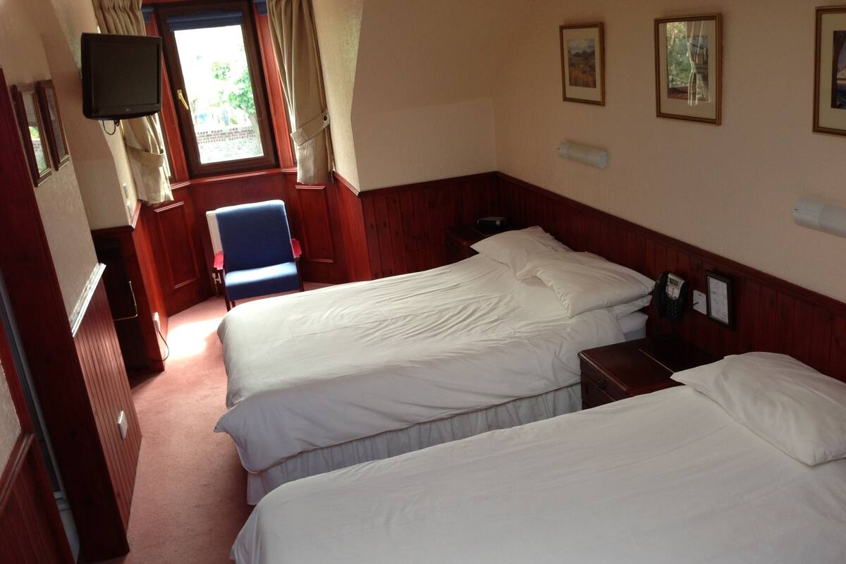 Ein Hotelzimmer mit zwei Einzelbetten, einem Sessel, einem Schreibtisch mit Telefon und einem an der Wand befestigten Fernseher.