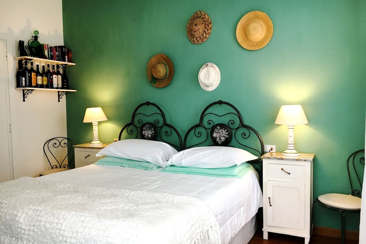 A neatly made bed with white linens, a green wall, and a shelf with books and bottles.