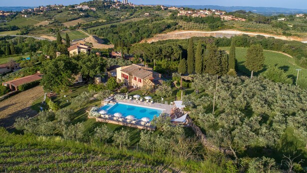 A villa with a swimming pool surrounded by lush greenery and vineyards, overlooking a hilly town with historic buildings. - Podere Bellavista (San Gimignano)