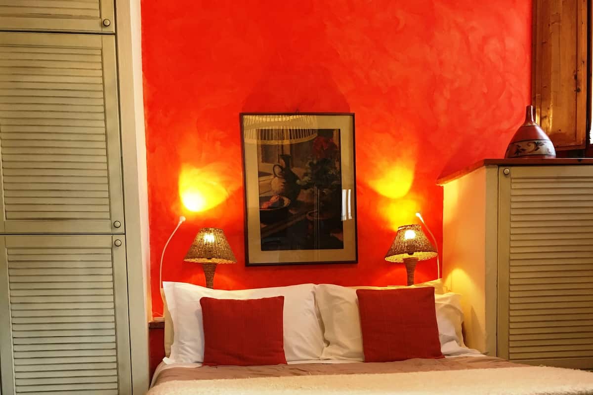 A bed with a grey and white duvet, two red pillows, and a white pillow. There are two bedside lamps with woven shades. A framed picture hangs on the wall above the bed.