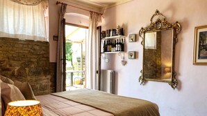 A bedroom with a stone wall, a bed with a floral-patterned skirt, a bedside lamp, a mirror, and a shelf with wine bottles. - Podere Bellavista (San Gimignano)