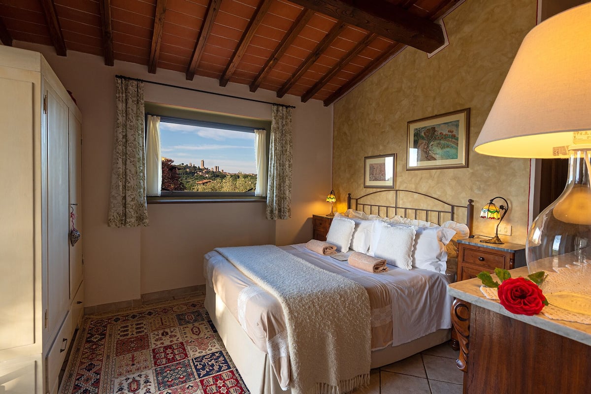 A bedroom with a bed, bedside lamps, a wooden headboard, a window with curtains, a patterned rug, and a red rose on the nightstand.