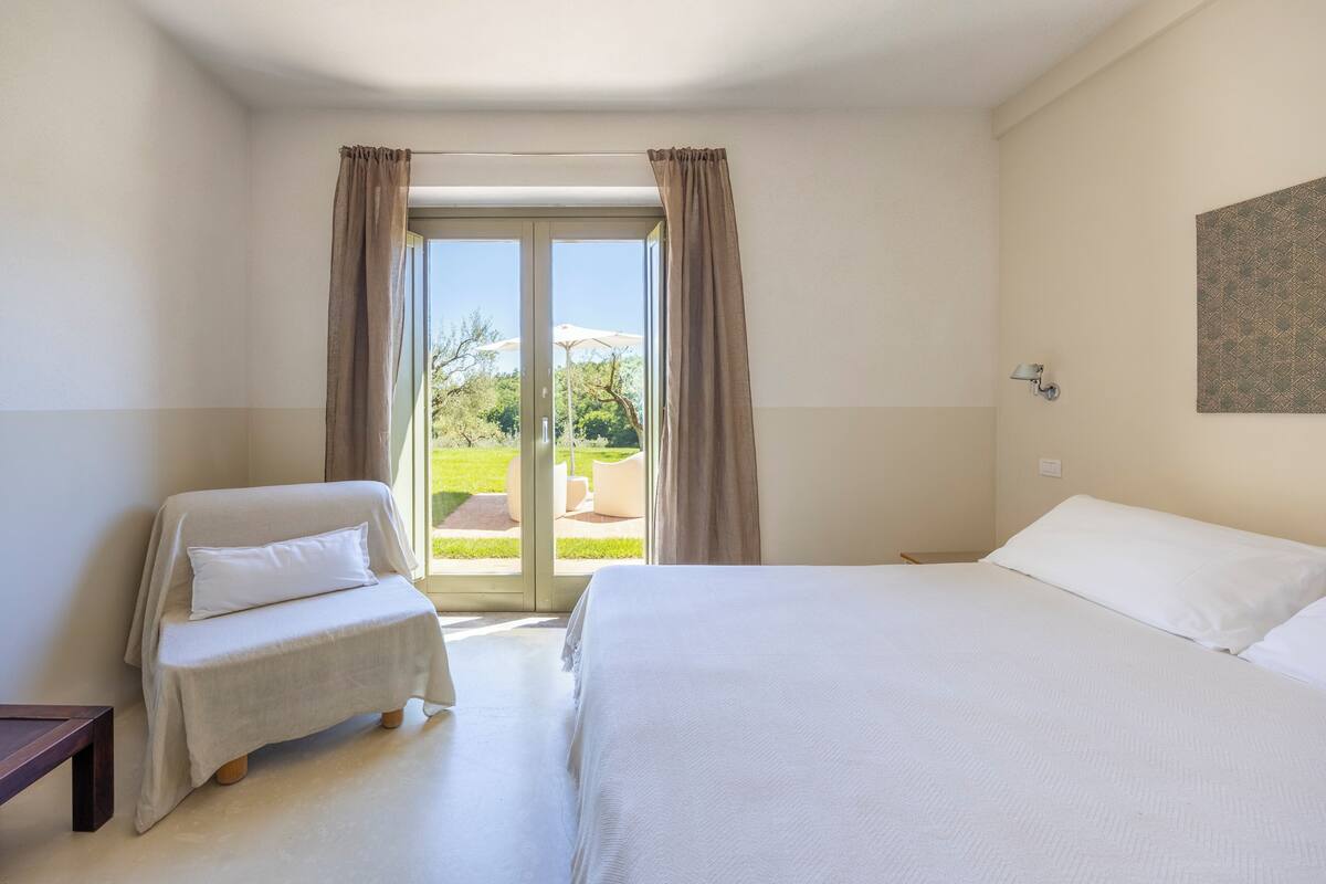 Un dormitorio con cama, una silla, un ventanal y una puerta que da a un espacio exterior.